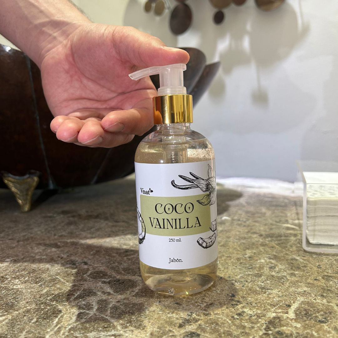 Jabón de Coco Vainilla de 250ml con una mano apunto de ser utilizado en un baño elegante con un lavamanos y toallas secadoras.