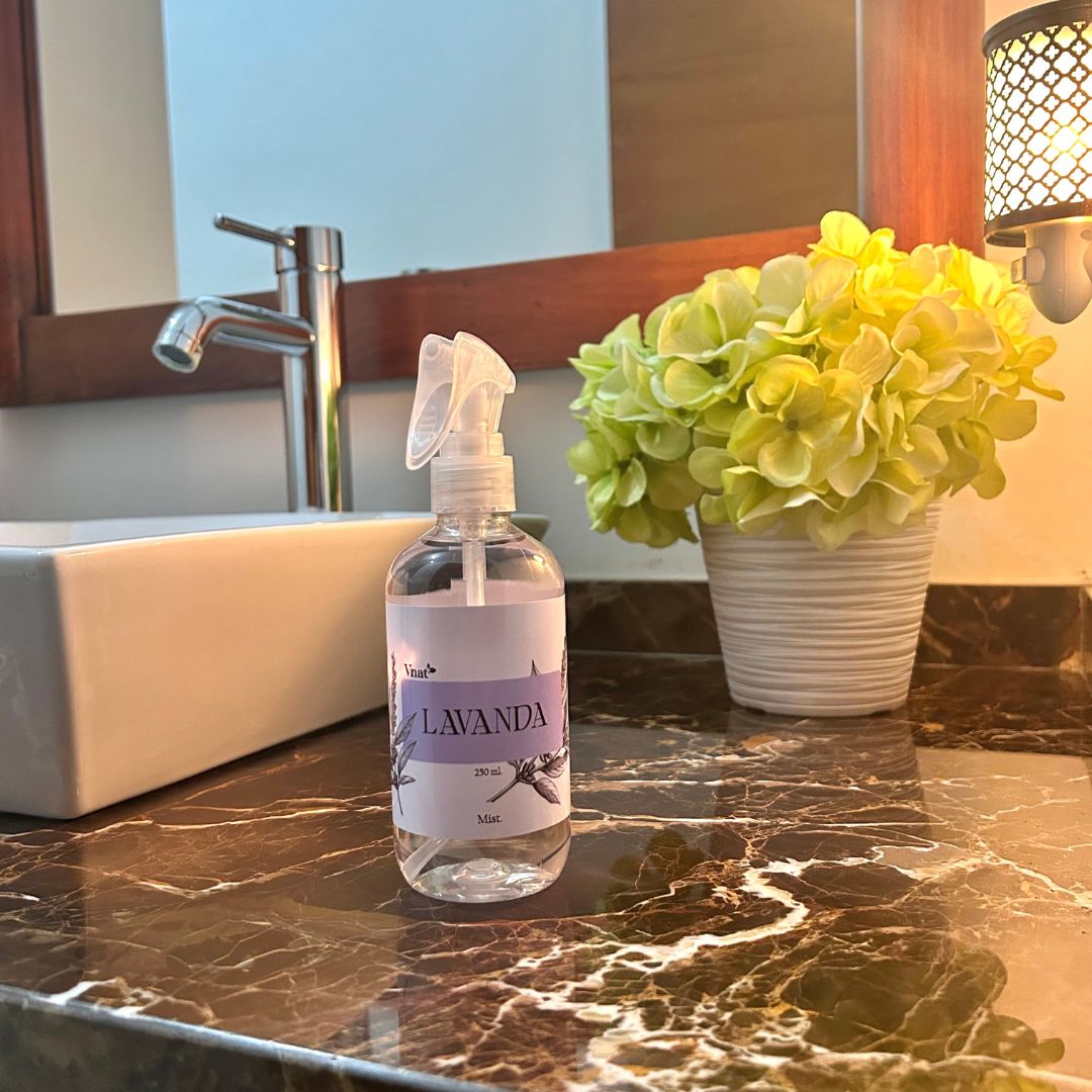 Mist aromatizante de lavanda de 250ml en un baño con luz tenue indirecta, junto a un lavamanos frente a un espejo y un adorno de plantas verdes.