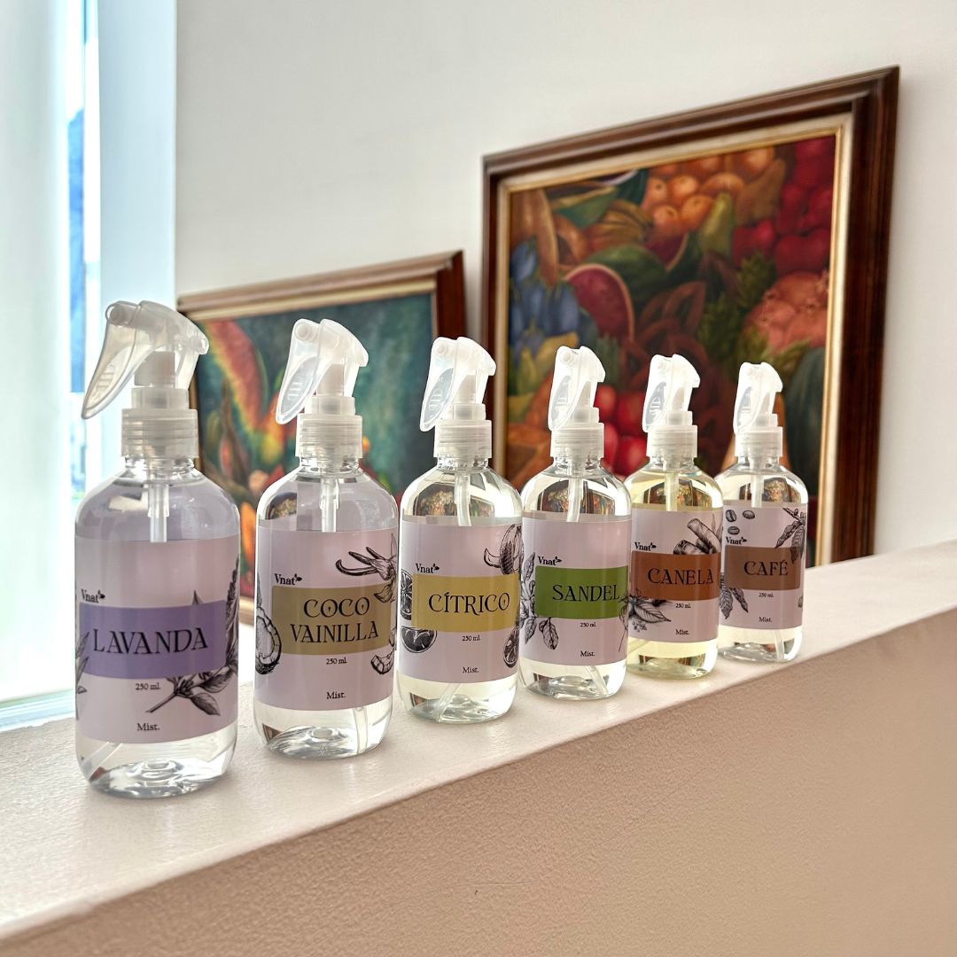 Mists aromatizantes de 250ml de varios olores (cítrico, canela, café, lavanda, coco vainilla, sándalo) en una barda con dos cuadros de pintura y un ventanal al fondo.