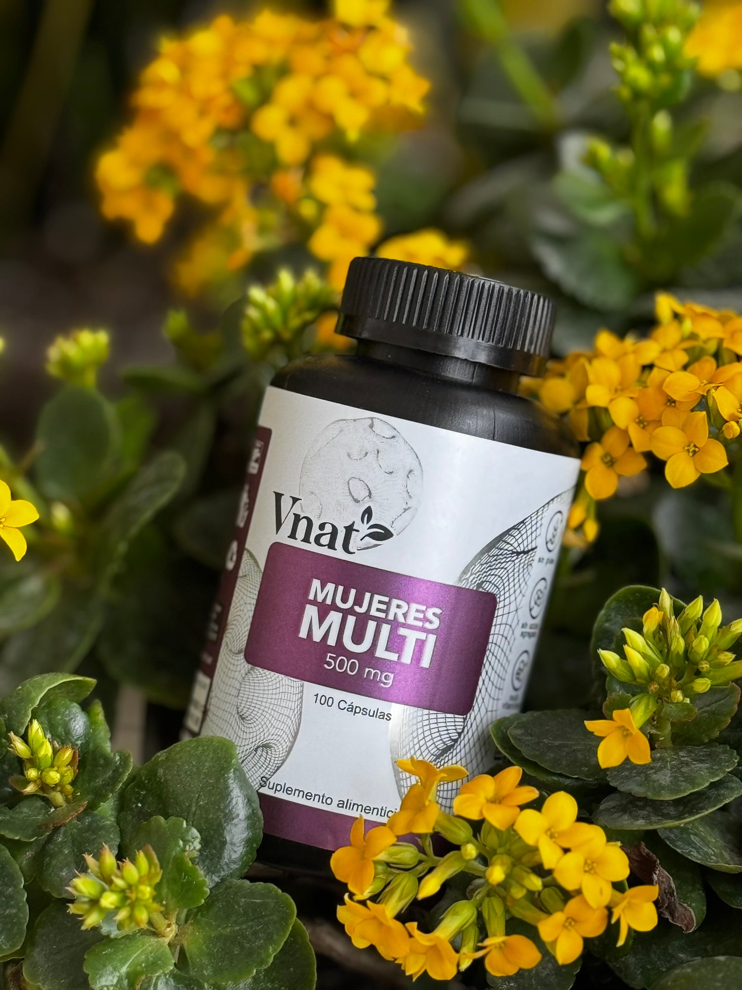 Frasco de suplemento Mujeres Multi 500 mg de la marca Vnat con 100 cápsulas, colocado entre flores amarillas y hojas verdes en un jardín.