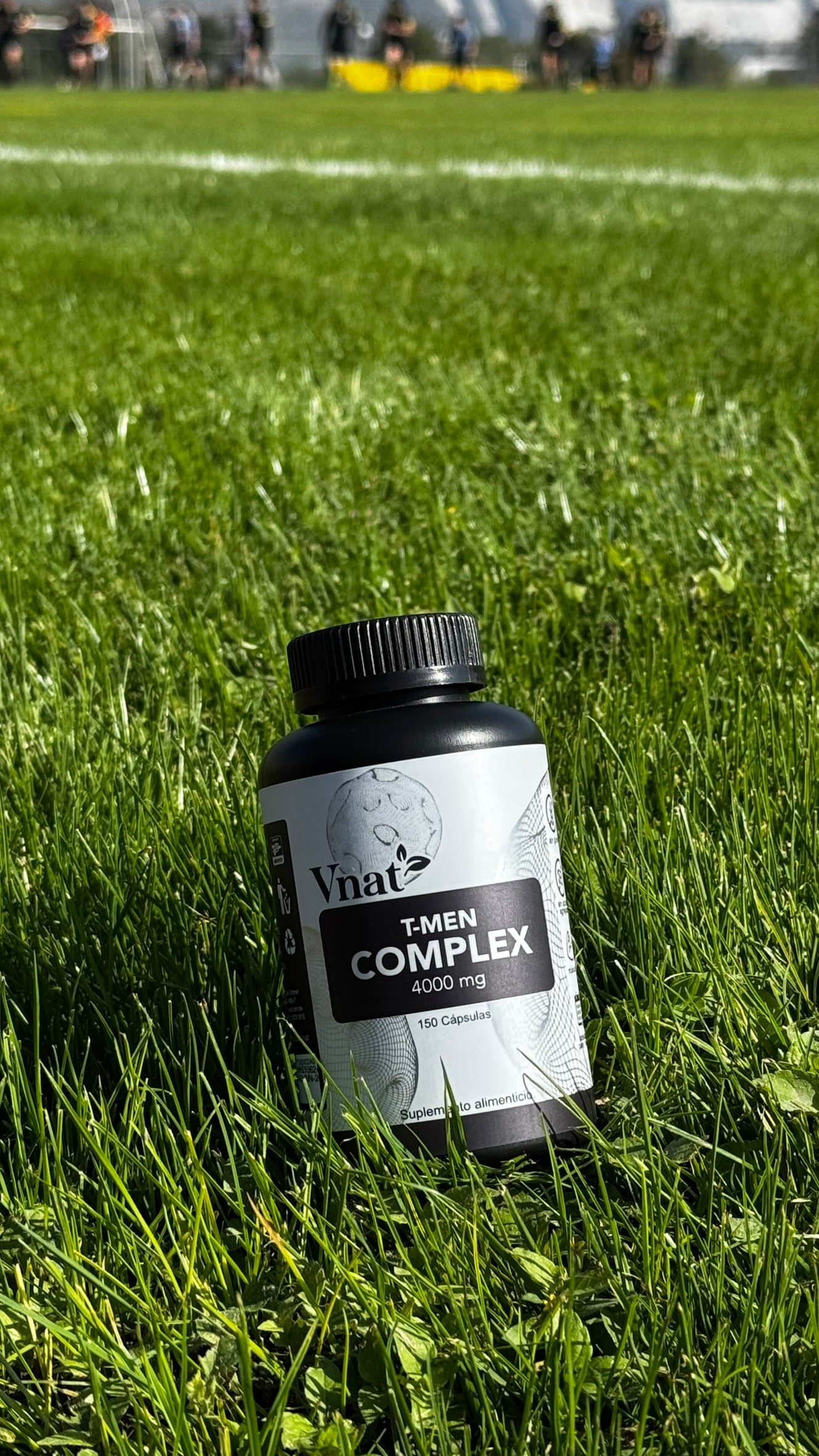 Frasco de suplemento T-Men Complex 4000 mg de la marca Vnat, con 150 cápsulas, colocado sobre césped verde con jugadores de fútbol desenfocados en el fondo.